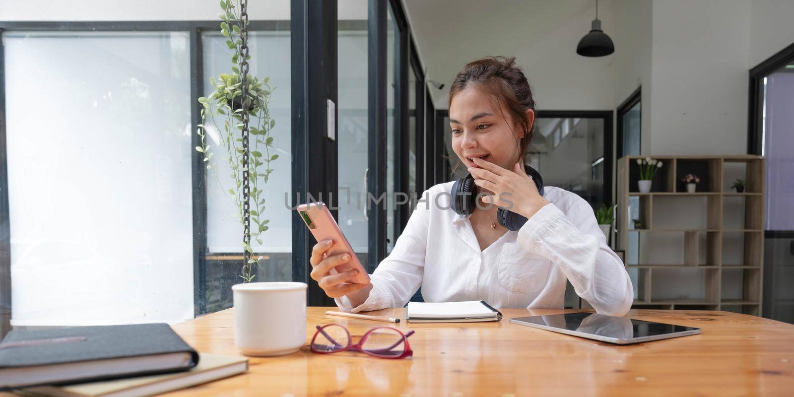 Happy young asian teen girl sit on sofa at home holding phone looking at screen waving hand video calling distance friend online in mobile chat app using smartphone videochat application. by wichayada