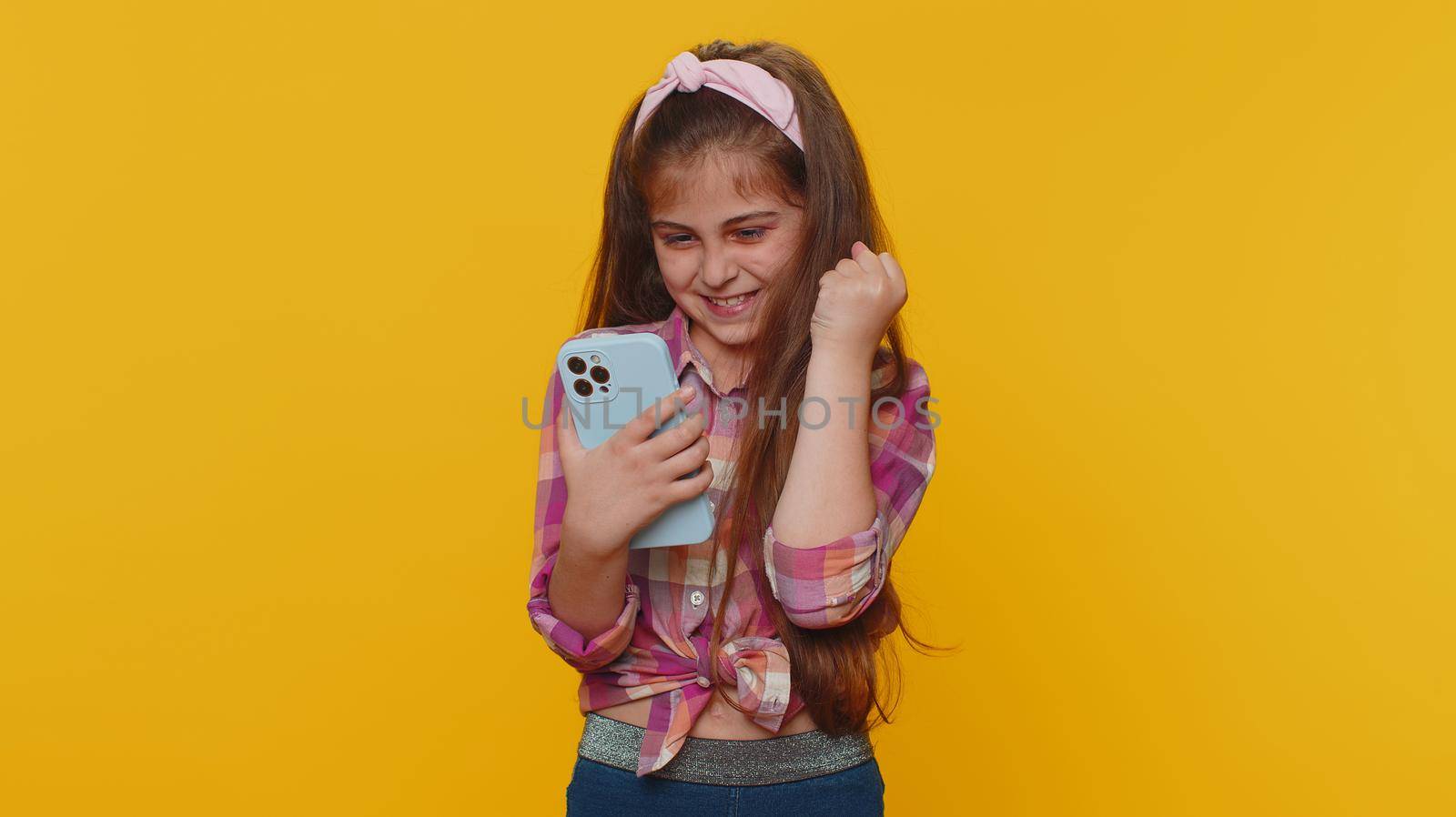 Young preteen child girl kid use mobile smartphone typing browsing say Wow yes found out great big win good news celebrate doing winner gesture. Little toddler children on studio yellow background