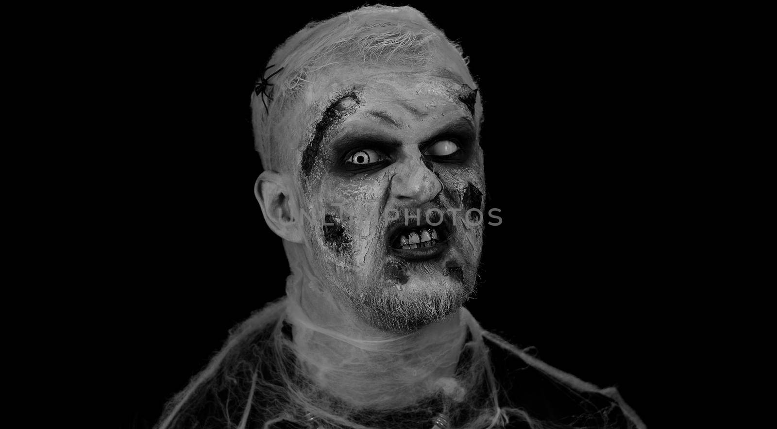 Frightening man with Halloween zombie bloody wounded makeup, trying to scare, face expressions. Horror theme. Sinister undead guy isolated on studio black background. Voodoo rituals. Fashion body art