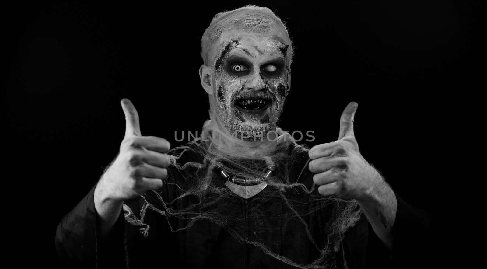Ugly man with scary Halloween zombie makeup in costume raises thumbs up agrees with something or gives positive reply recommends advertisement likes good. Dead guy with wounded bloody scars face