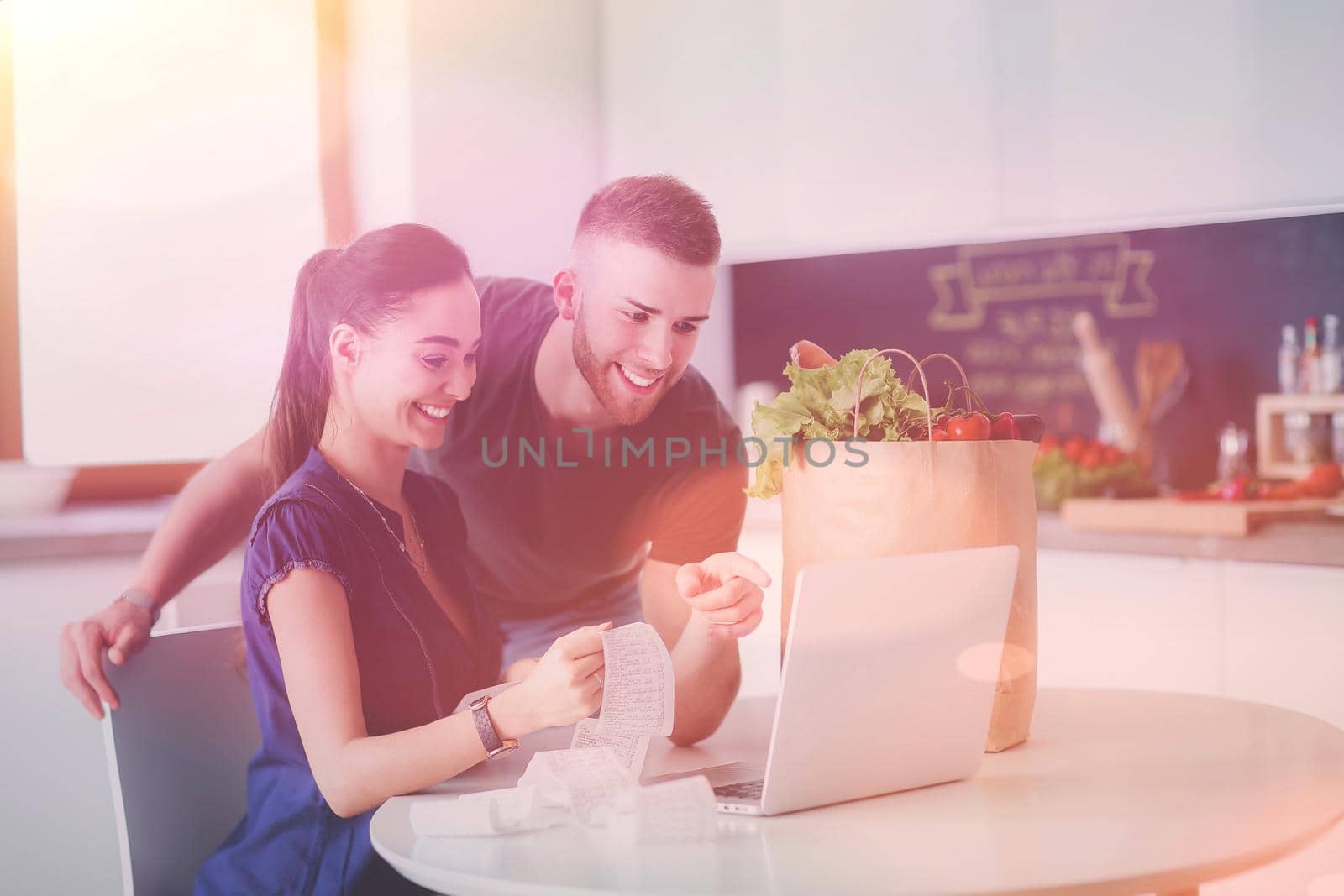 Young man cutting vegetables and woman standing with laptop in the kitchen by lenets