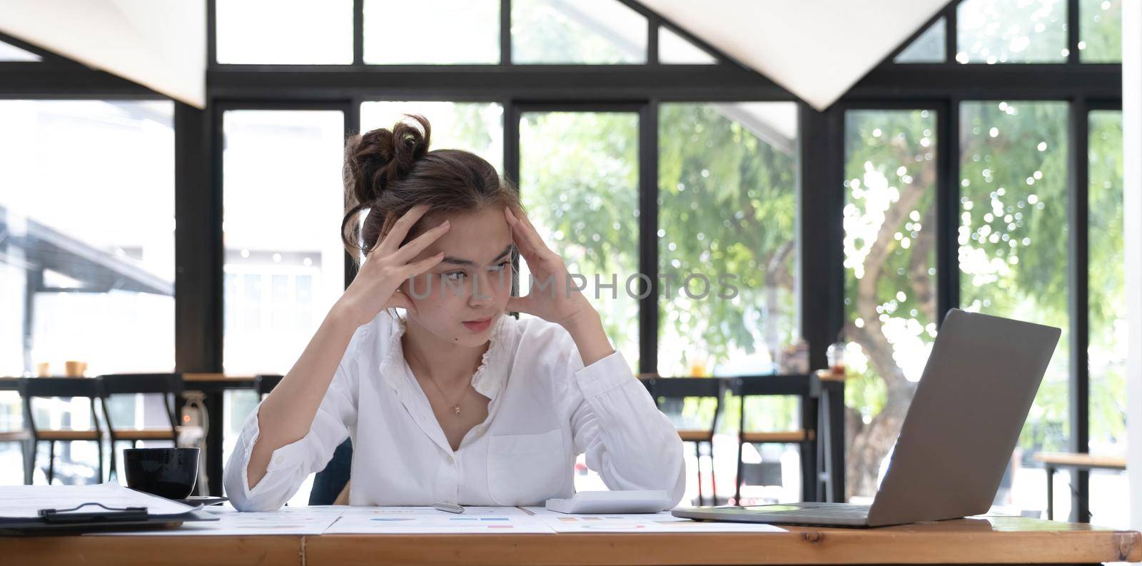 Photo of a tired woman keeps a hand on her head at the wooden working desk after finishing work hard..