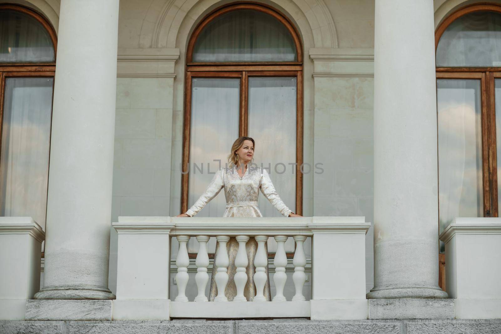 a beautiful mature woman in a long white dress stands on the balcony and looks into the distance.