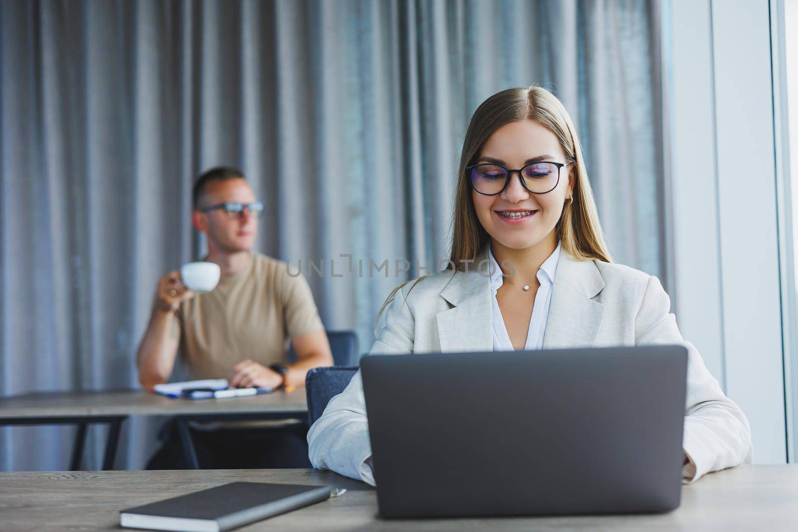 Attractive young woman in glasses sits at a table with a laptop in a coworking space and discusses a project plan with a colleague. Workflow in the office.