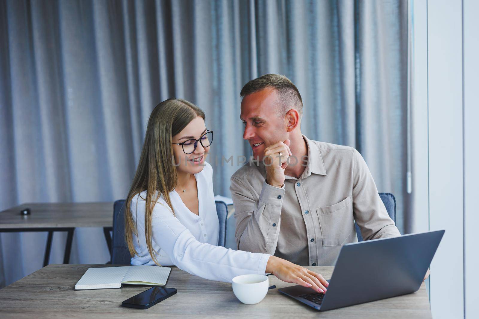 Businessmen are discussing while working with laptop in the office. Focused business people cooperating in a modern workspace. Two young businessmen are sitting together at a table. by Dmitrytph