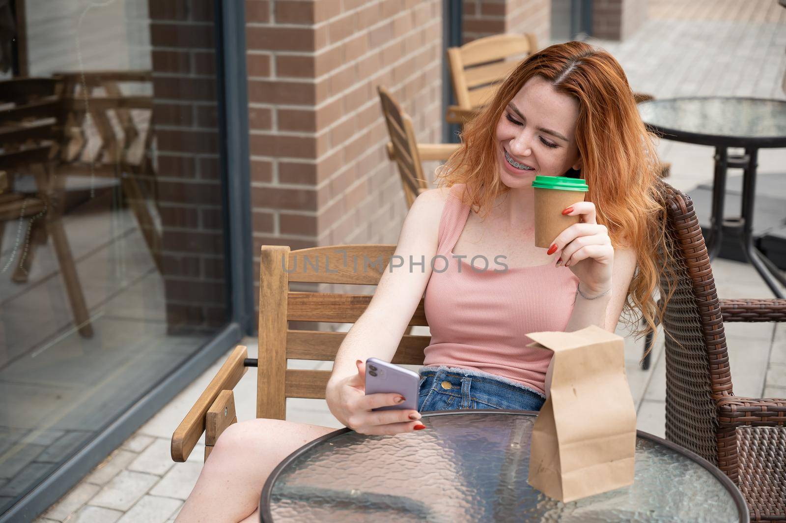 Young red-haired woman with braces on her teeth drinks coffee on a summer veranda
