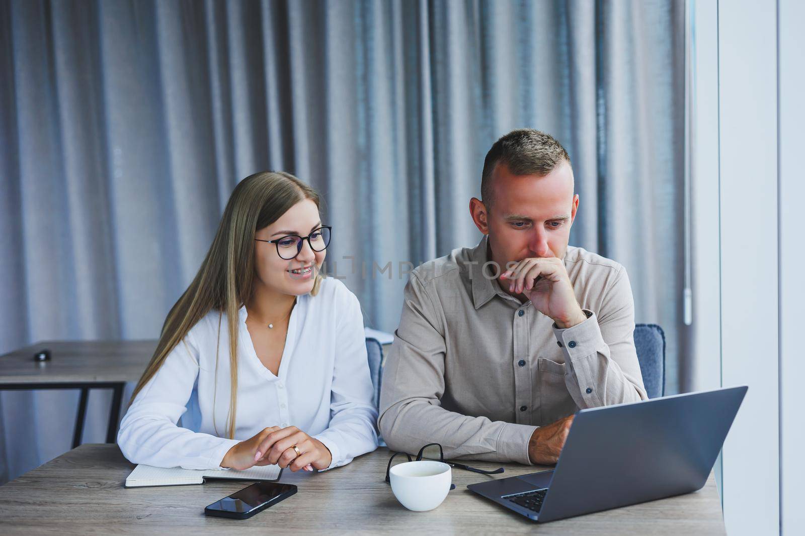 Businessmen are discussing while working with laptop in the office. Focused business people cooperating in a modern workspace. Two young businessmen are sitting together at a table.