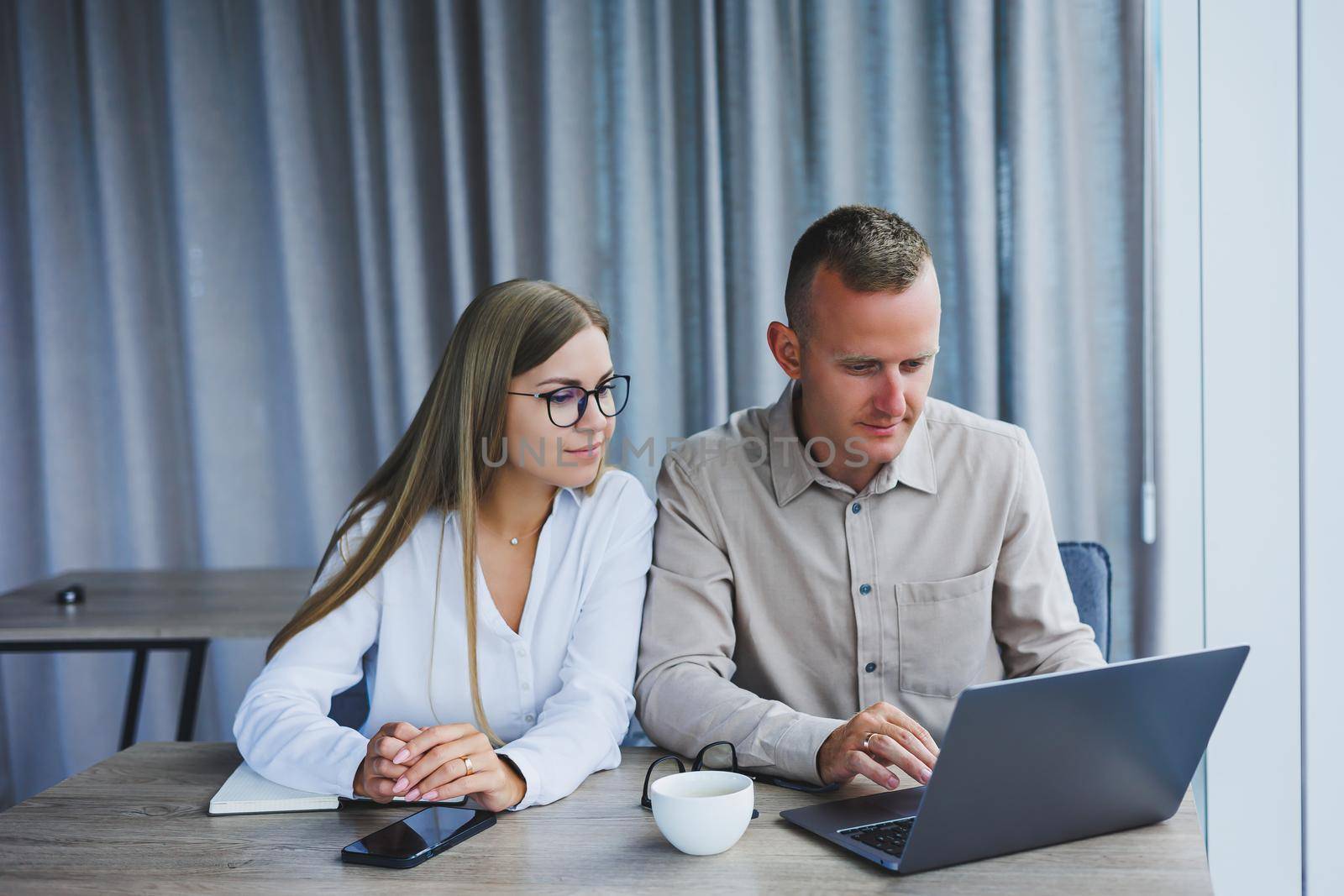 Businessmen are discussing while working with laptop in the office. Focused business people cooperating in a modern workspace. Two young businessmen are sitting together at a table.