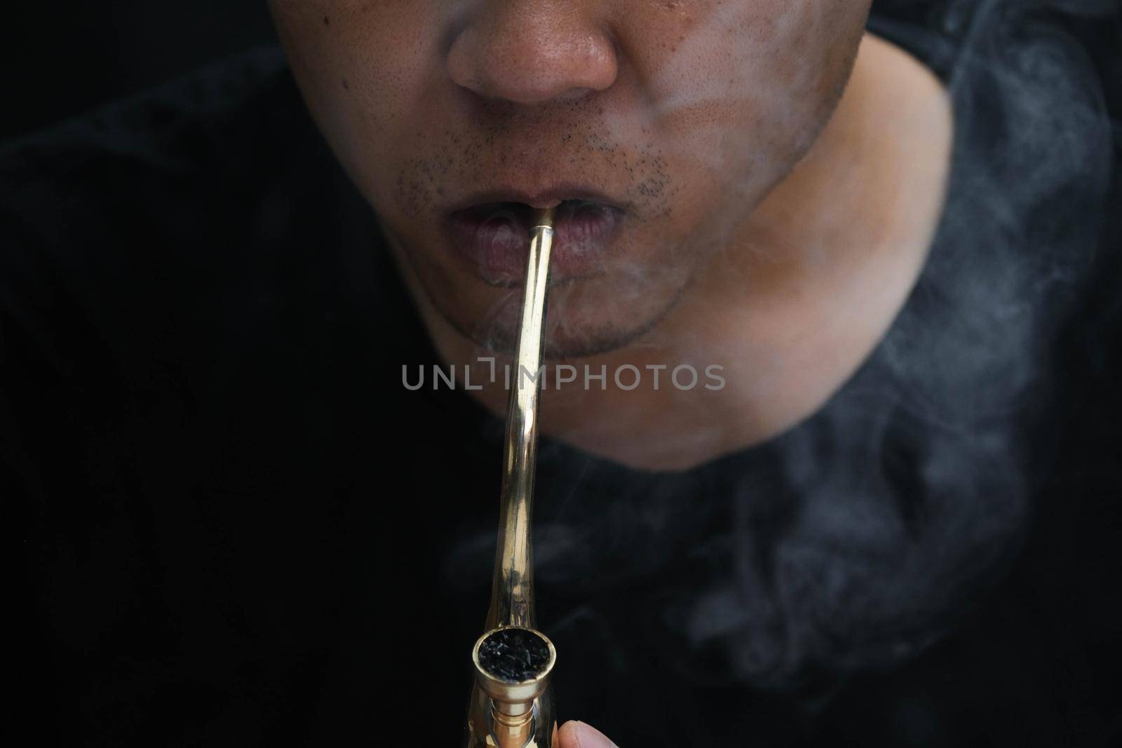 Asian man smokes marijuana from a pipe at home. Studio shoot with model simulating smoking pot with a pipe in a dark background. Cannabis legalisation.