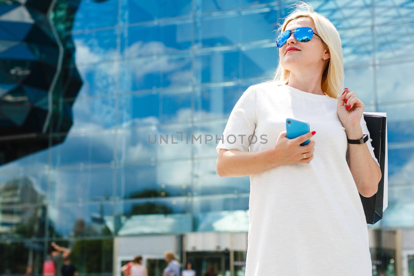 Beautiful girl is holding shopping bags, using a smart phone and smiling while doing shopping in the city by Andelov13