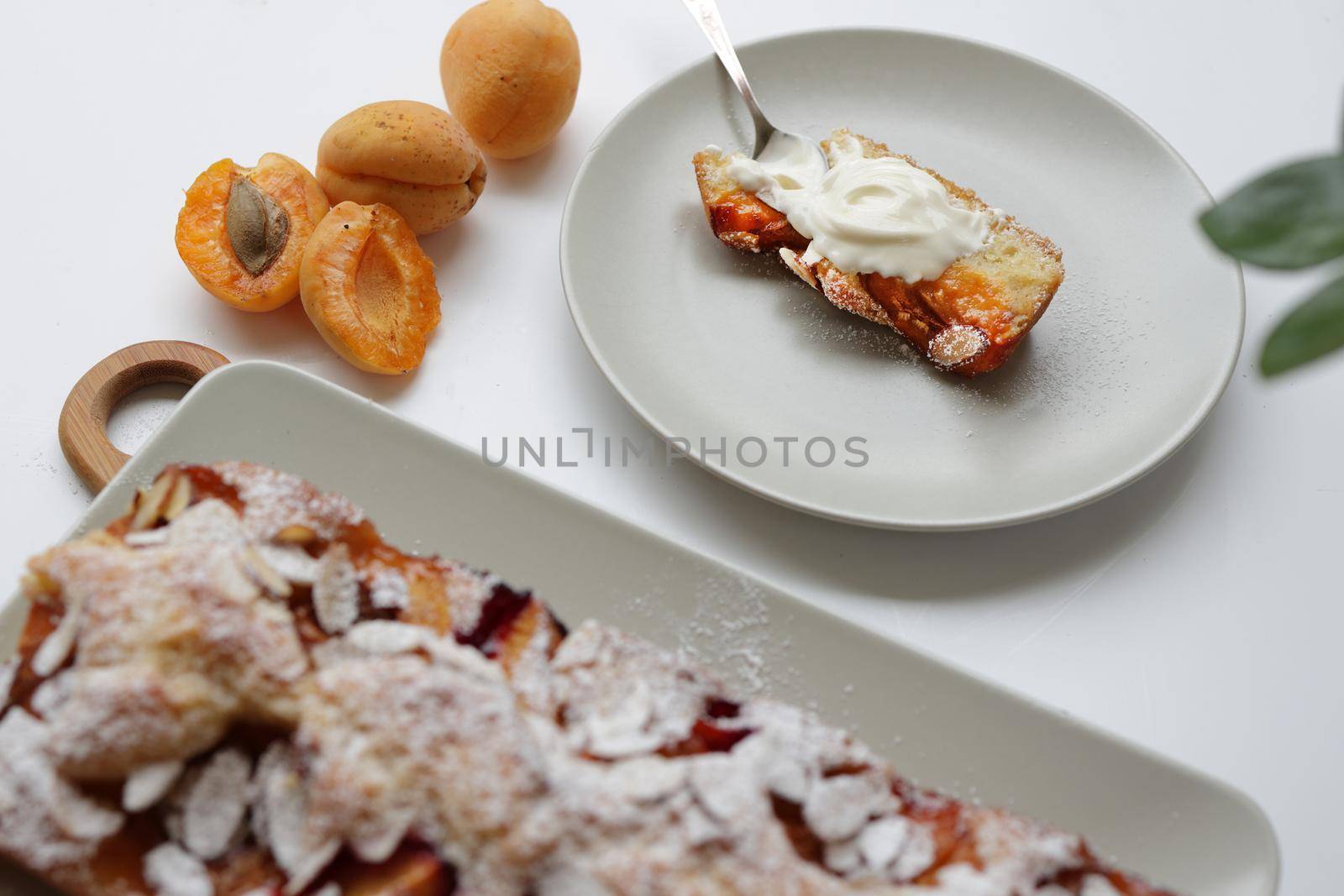 rectangular appetizing homemade apricot pie on a rectangular plate, on a wooden cutting board. Next to it is a round plate with a piece of cake and a spoonful of cream. Daylight, white background