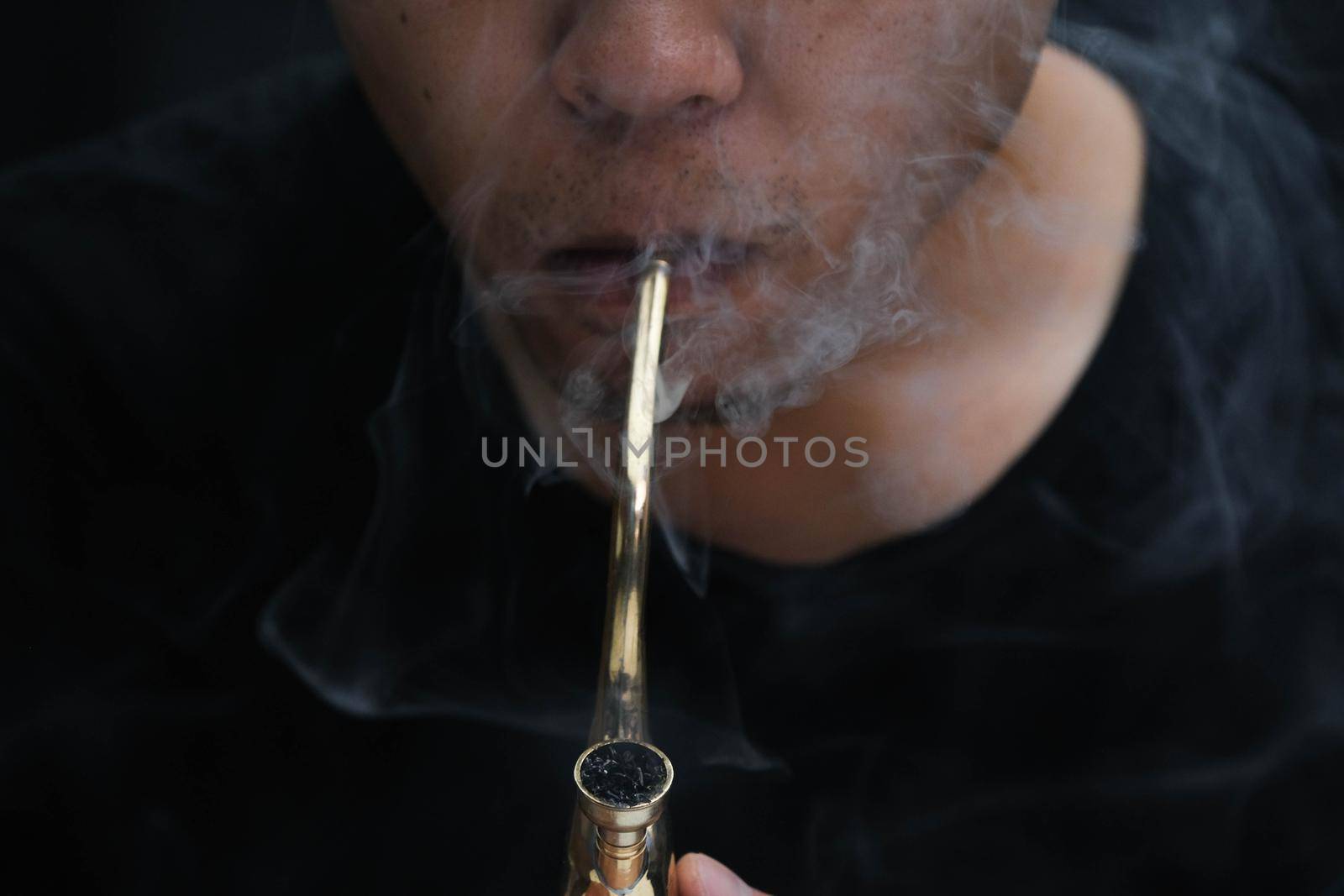 Asian man smokes marijuana from a pipe at home. Studio shoot with model simulating smoking pot with a pipe in a dark background. Cannabis legalisation. by TEERASAK