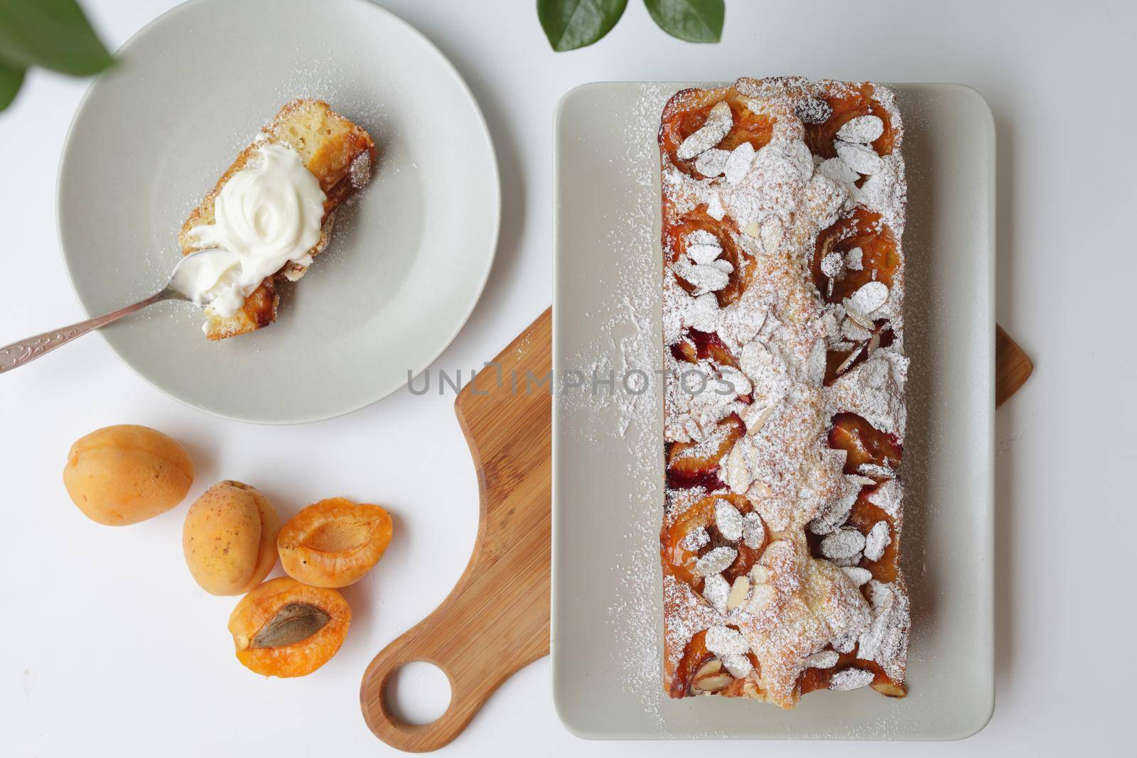 rectangular appetizing homemade apricot pie on a rectangular plate, on a wooden cutting board. Next to it is a round plate with a piece of cake and a spoonful of cream. Daylight, white background, top view, flat lay