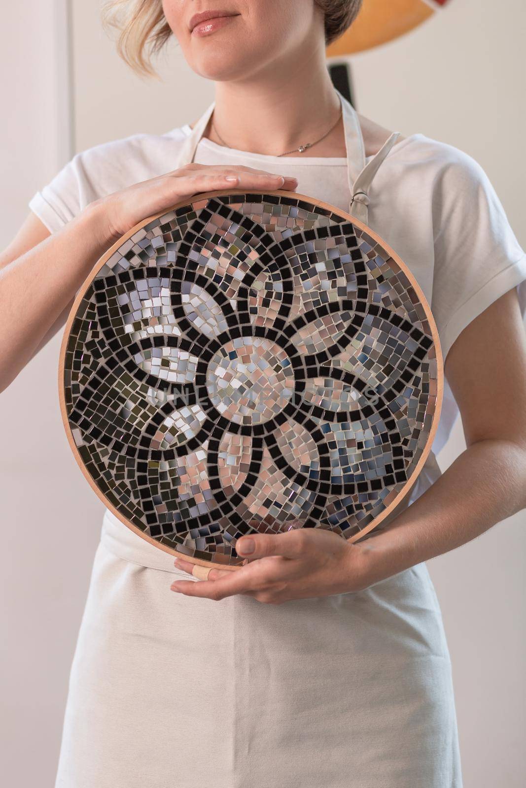 Young blonde woman in a long beige apron is standing in a small workshop and holding in hands mosaic plate. Close-up