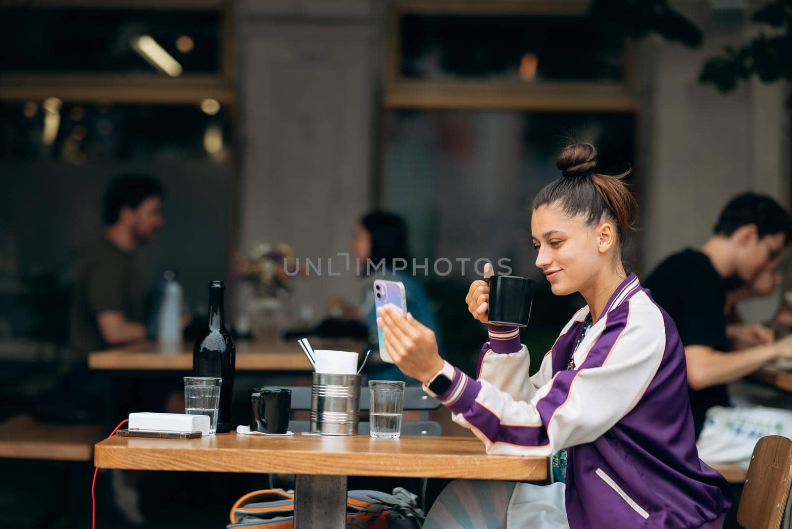 Young woman sitting outdoors on terrace in cafe at the table drinking beverage tea or coffee holding in hand smartphone