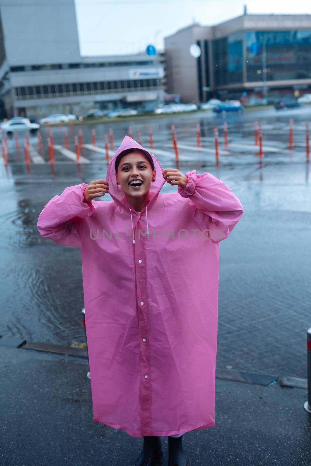 Young smiling woman with a pink raincoat on the street while enjoying a walk through the city on a rainy day.