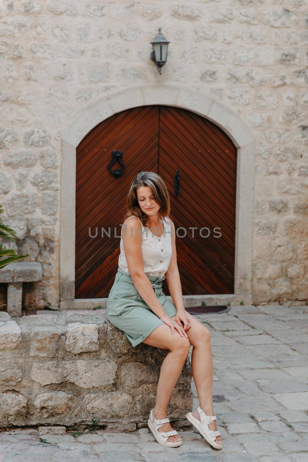 Girl Tourist Resting in the Ancient Narrow Street On A Beautiful Summer Day In MEDITERRANEAN MEDIEVAL CITY, OLD TOWN KOTOR, MONTENEGRO. Young Beautiful Cheerful Woman Walking On Old Street