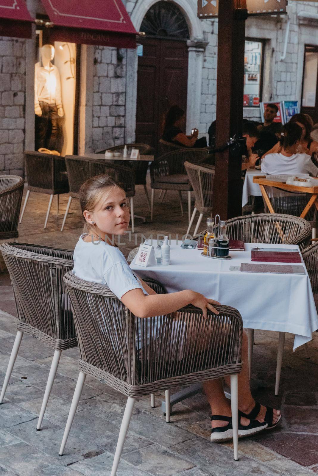 Girl Tourist Resting in the Ancient Narrow Street On A Beautiful Summer Day In MEDITERRANEAN MEDIEVAL CITY, OLD TOWN KOTOR, MONTENEGRO. Young Beautiful Cheerful Woman Walking On Old Street