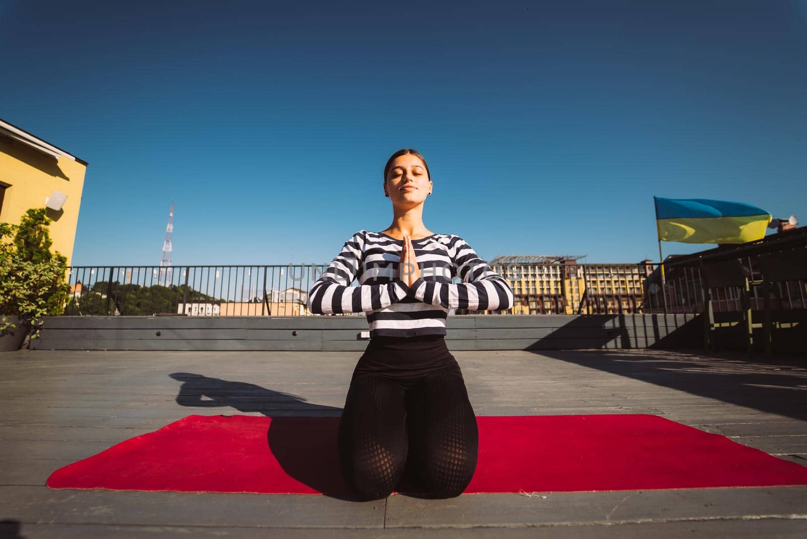 Young sporty happy woman in sportswear doing stretching exercises on yoga mat on house roof in early morning