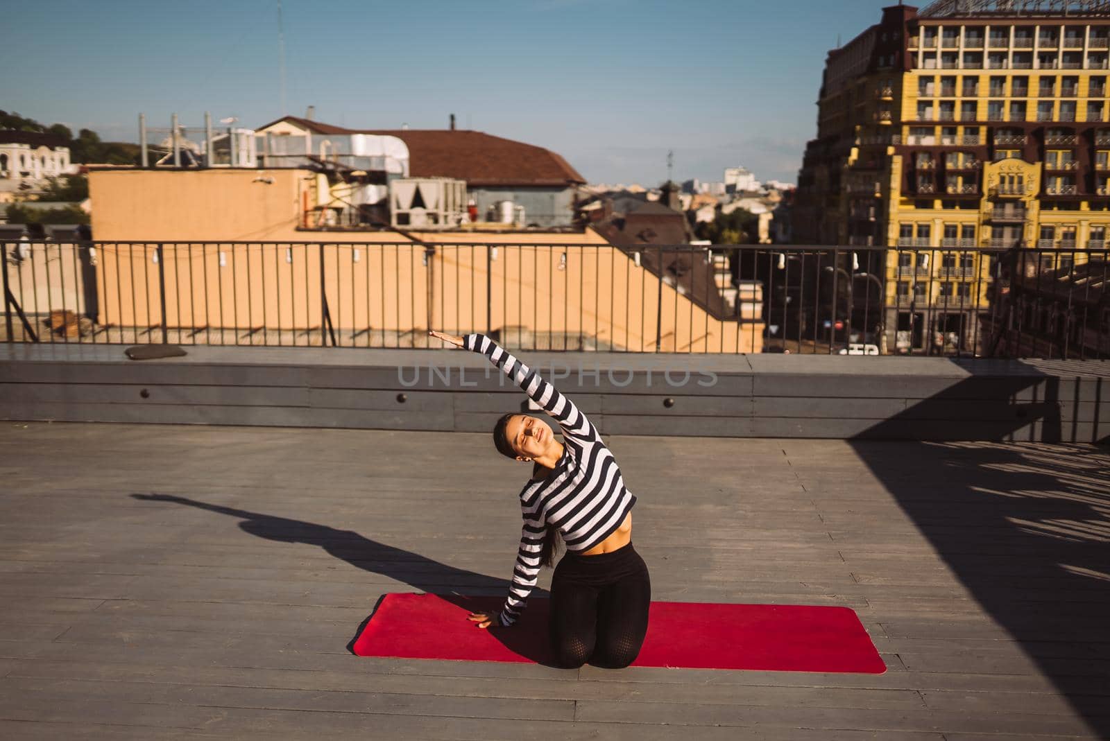 Young sporty happy woman in sportswear doing stretching exercises on yoga mat on house roof in early morning