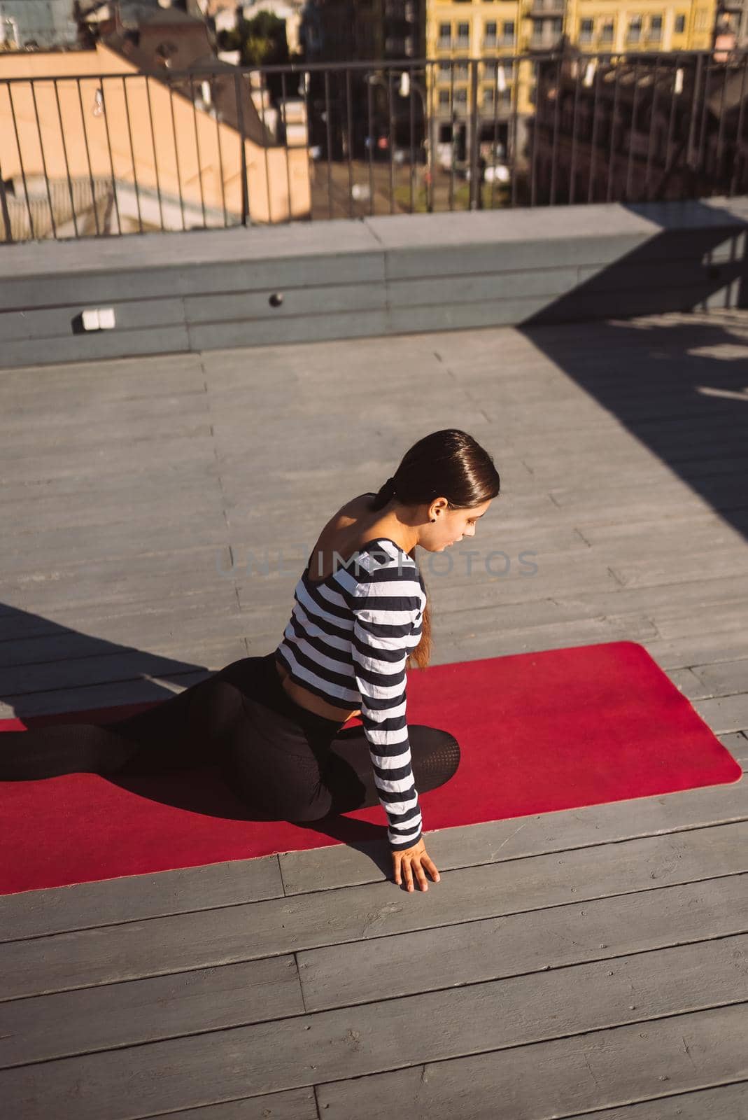 Young sporty happy woman in sportswear doing stretching exercises on yoga mat on house roof in early morning