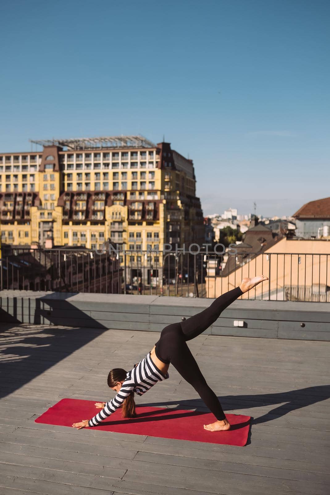 Young sporty happy woman in sportswear doing stretching exercises on yoga mat on house roof in early morning