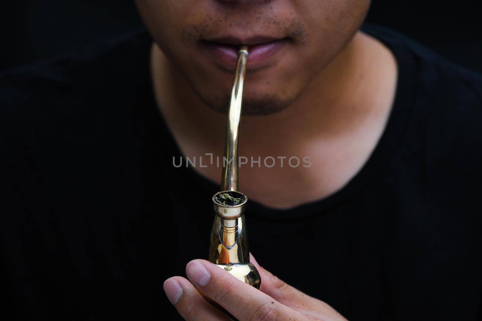 Asian man smokes marijuana from a pipe at home. Studio shoot with model simulating smoking pot with a pipe in a dark background. Cannabis legalisation. by TEERASAK