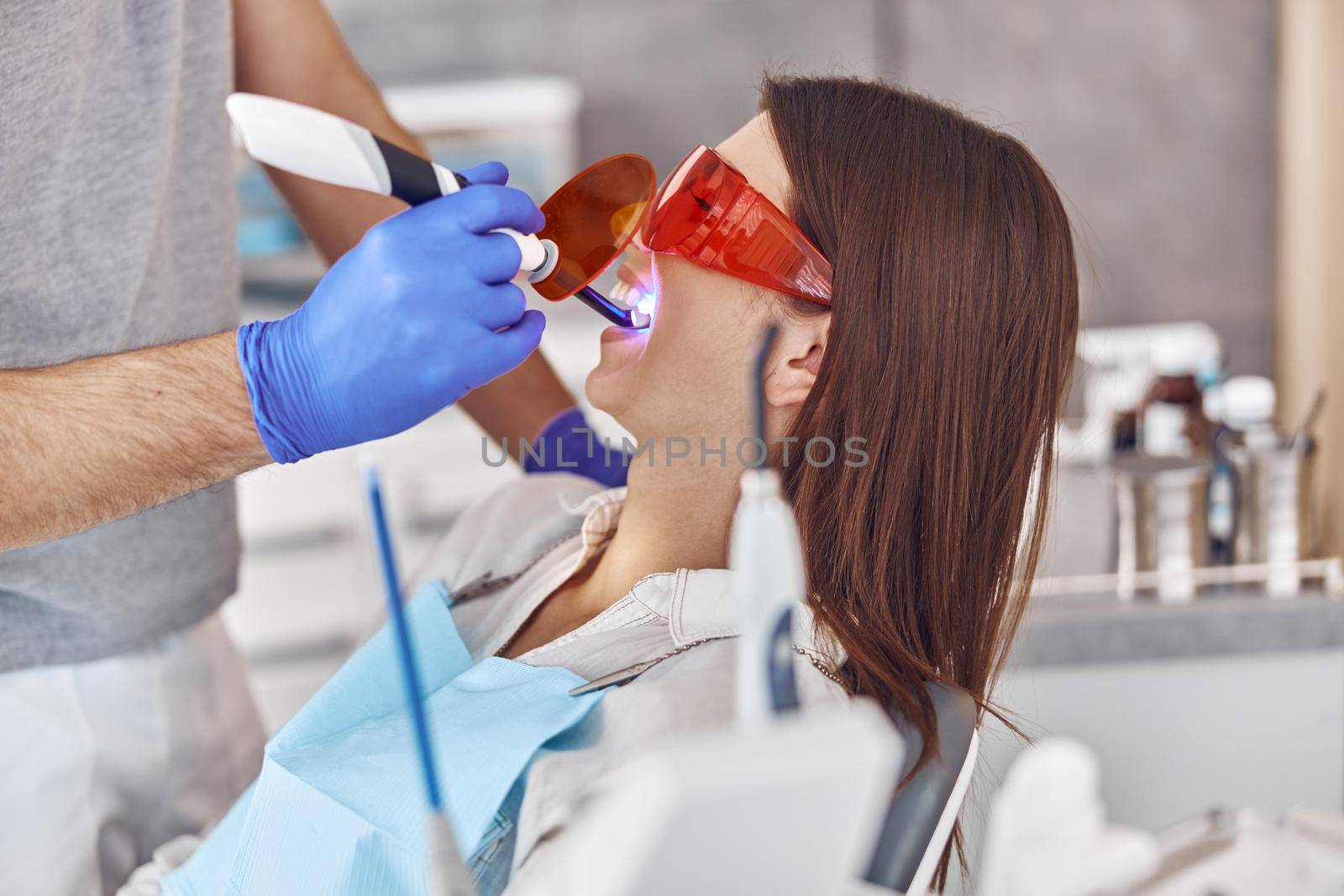 Professional male doctor uses ultraviolet lamp after making a filling on a tooth for joyful happy caucasian woman in modern dental cabinet by Yaroslav_astakhov