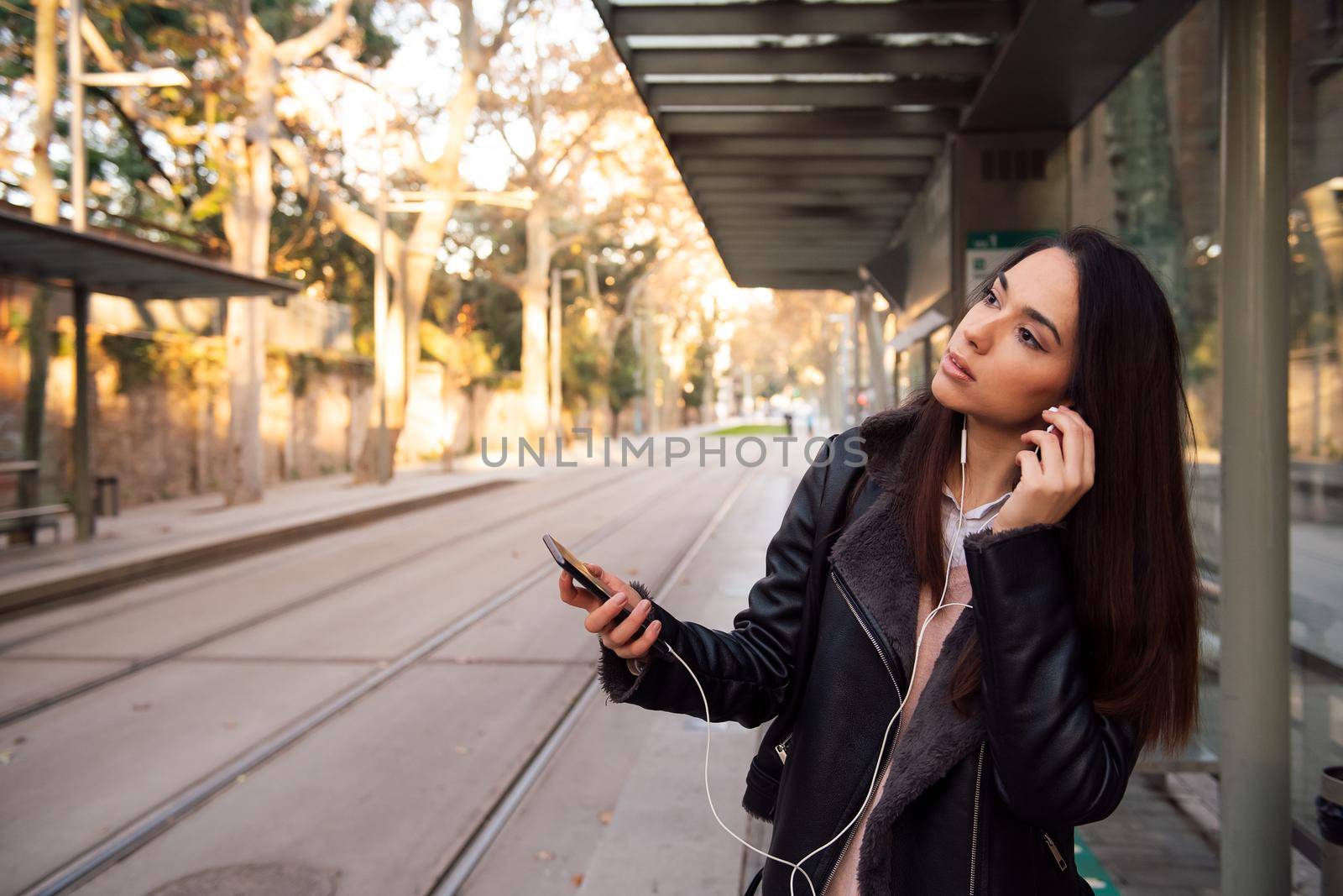 woman putting on her telephone earphones at the streetcar stop, concept of technology and communication, copyspace for text