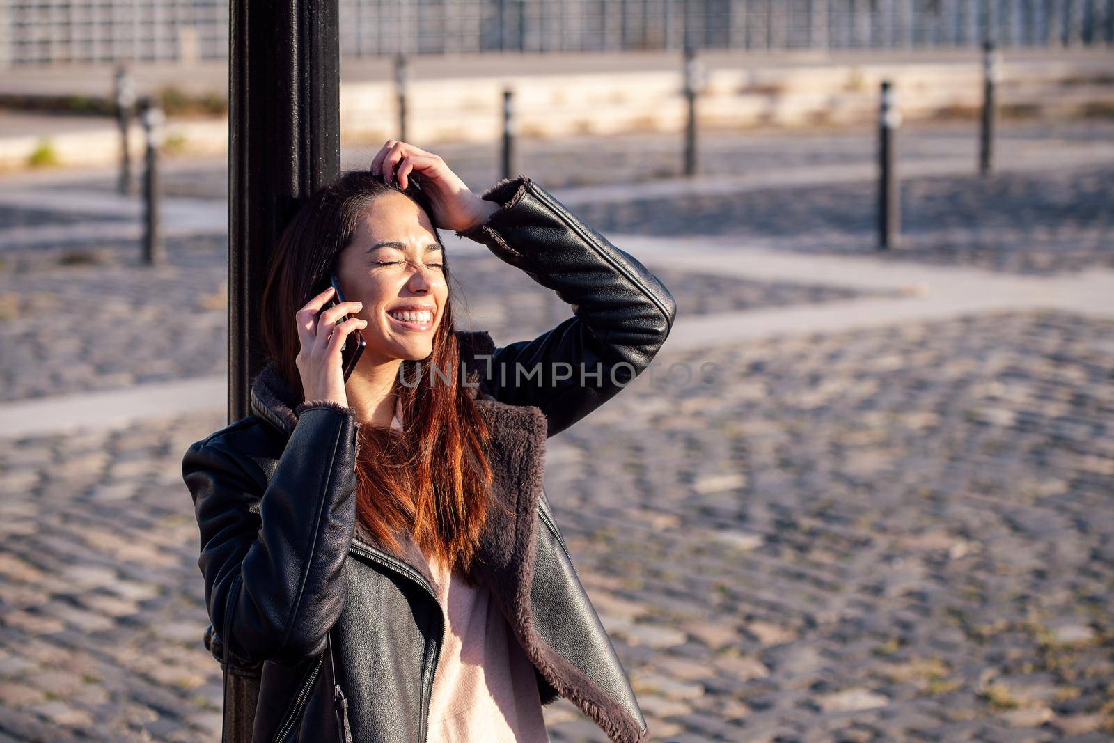 young woman laughing and talking on the phone leaning against a lamppost, concept of technology and urban lifestyle, copyspace for text
