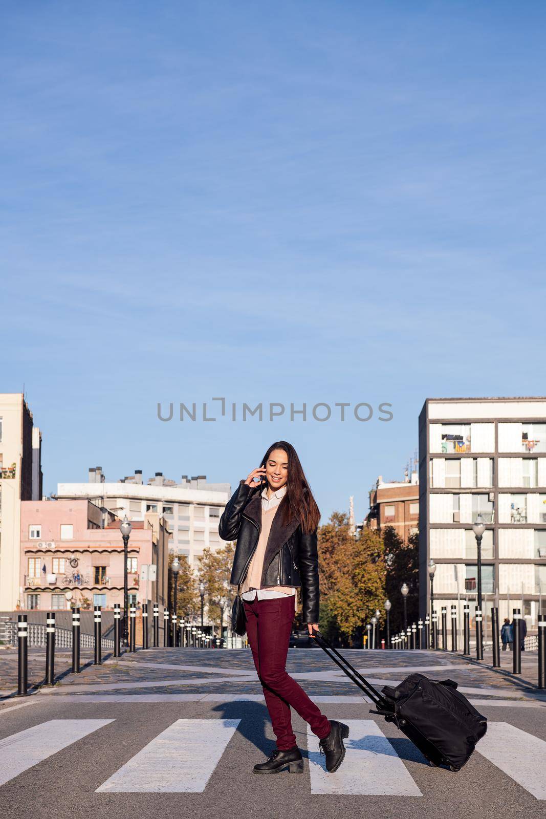 vertical photo of a young woman pulling suitcase talking by phone while crossing the street at a pedestrian crosswalk, concept of travel and urban lifestyle, copyspace for text