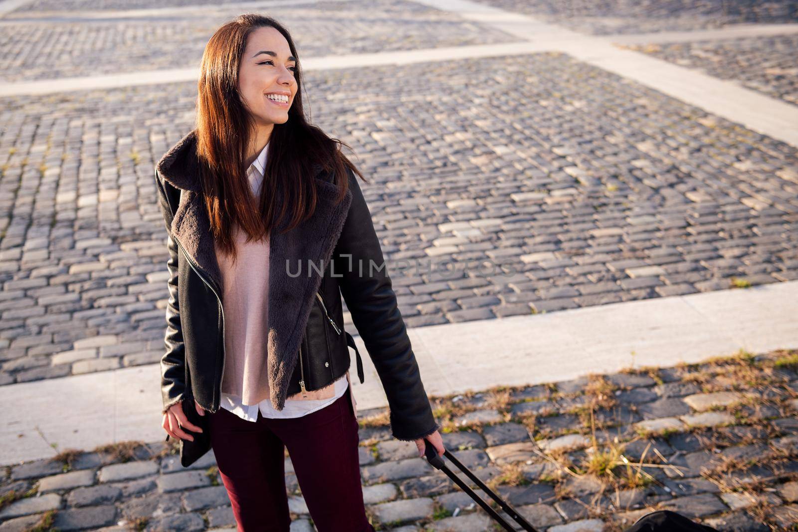 smiling young woman with trolley suitcase in a cobblestone street, concept of travel and urban lifestyle, copyspace for text