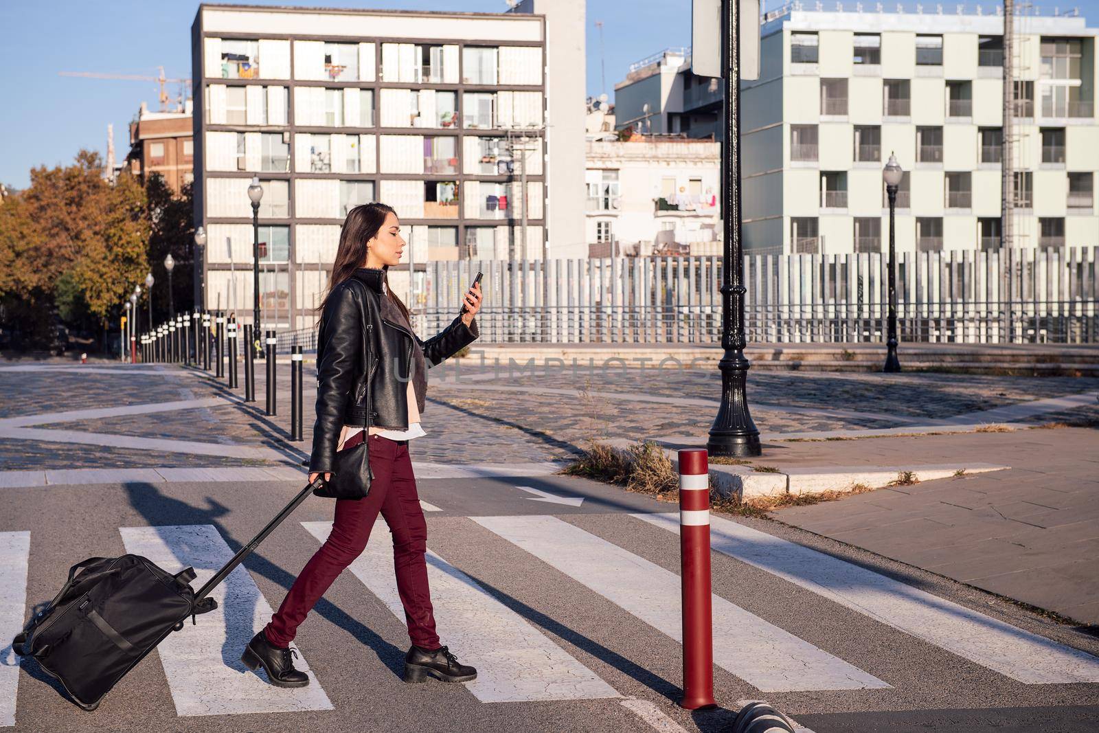 woman with trolley suitcase consulting phone while crossing the street at a pedestrian crosswalk, concept of travel and urban lifestyle