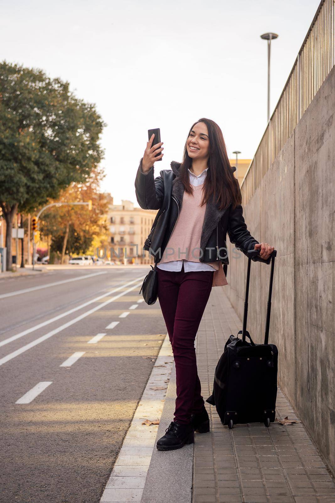 smiling young woman with suitcase waiting for a cab on the street while making a video call with her phone, concept of travel and technology, copyspace for text