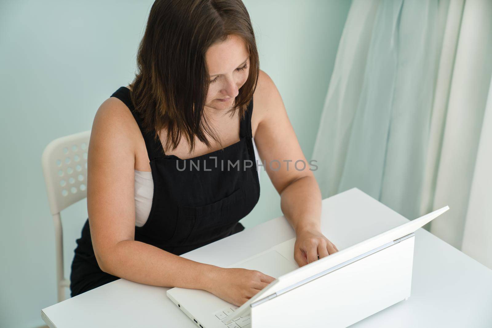 Young woman is sitting in front of her laptop transformer at desk in her house. Concept of freelance and distance education, side view