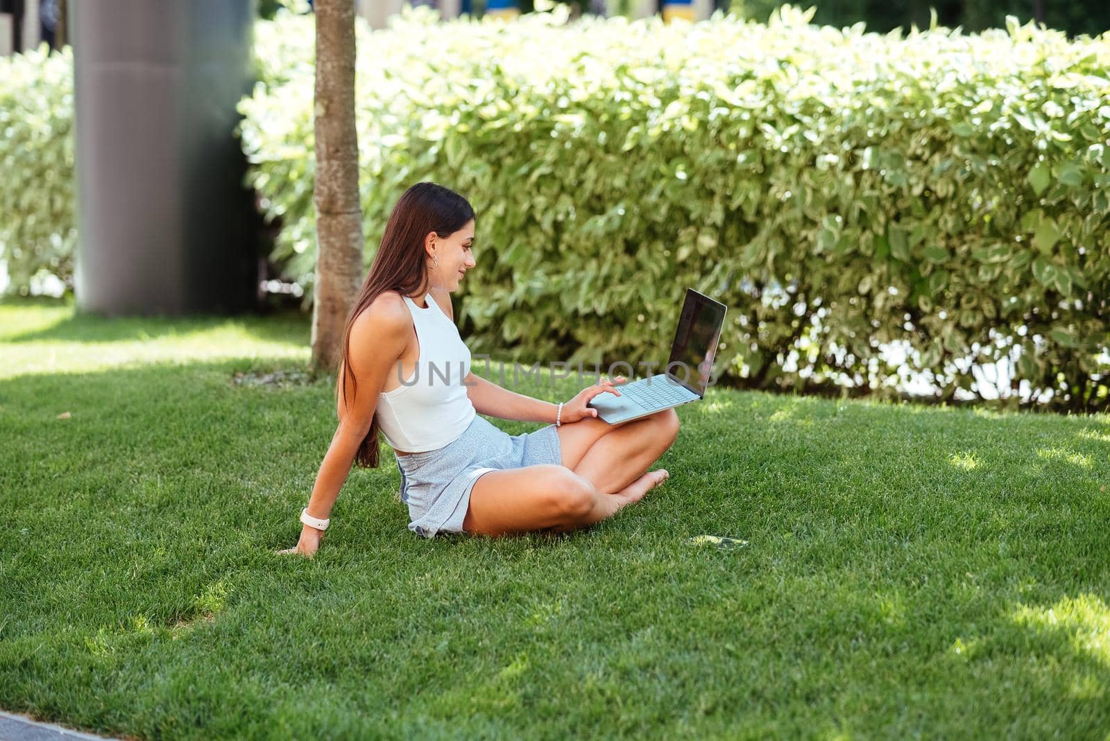 The concept of remote work. A young girl works with a laptop in the fresh air in the park, sitting on the lawn.