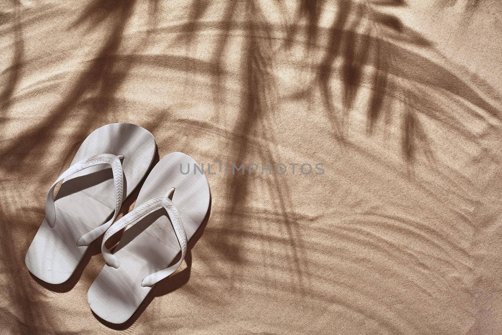Summer concept with a shadow of a tropical palm tree leaves, copyspace. White thongs are on a clean beach sand. Summertime lifestyle, objects in flat lay, top view arrangement.