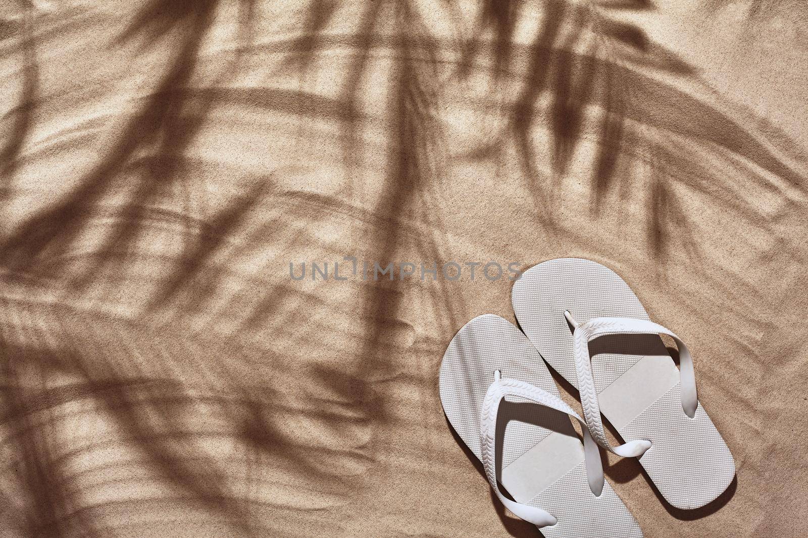 Summer concept with a shadow of a tropical palm tree leaves, copyspace. White thongs are on a perfect beach sand. Summertime lifestyle, objects in flat lay, top view arrangement.