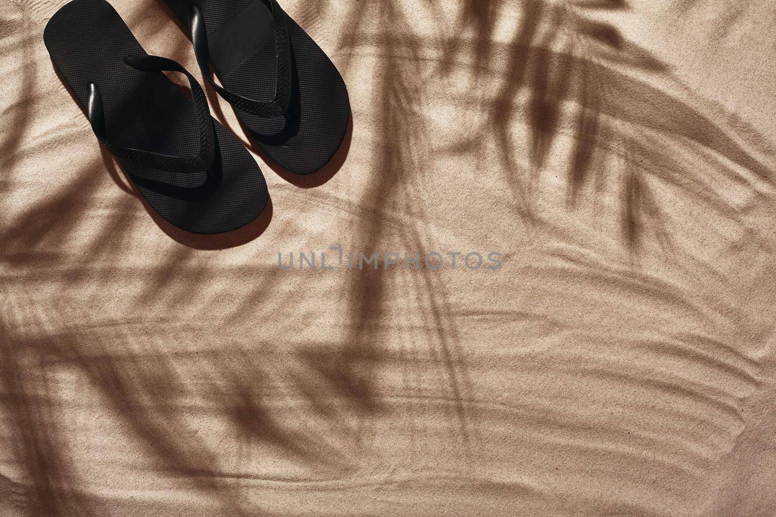 Summer concept with a shadow of a tropical palm tree leaves, copyspace. Black thongs are on a clean beach sand. Summertime lifestyle, objects in flat lay, top view arrangement.