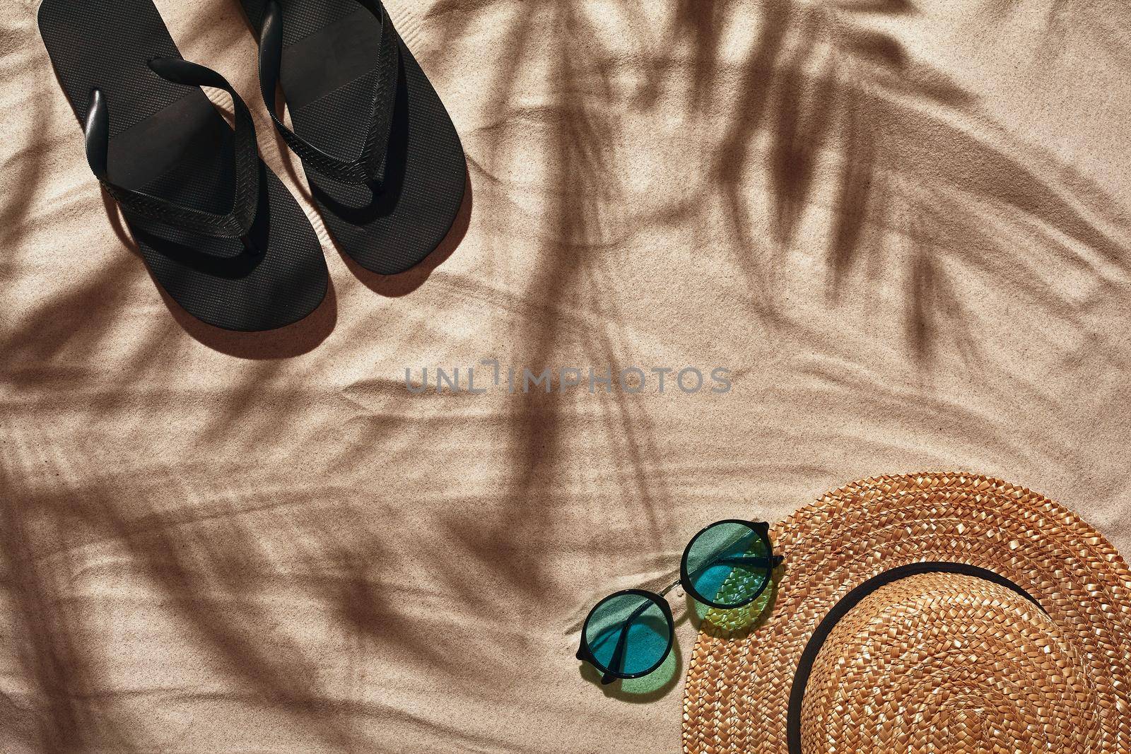 Summer concept with a shadow of a tropical palm tree leaves, copyspace. Traveler vacation accessories such as black thongs, sunglasses and a straw hat are laid out on a white beach sand. Summertime lifestyle, objects in flat lay, top view arrangement.