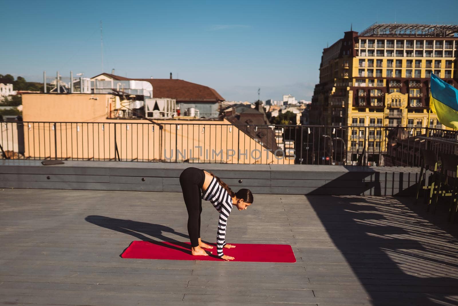 Young sporty happy woman in sportswear doing stretching exercises on yoga mat on house roof in early morning