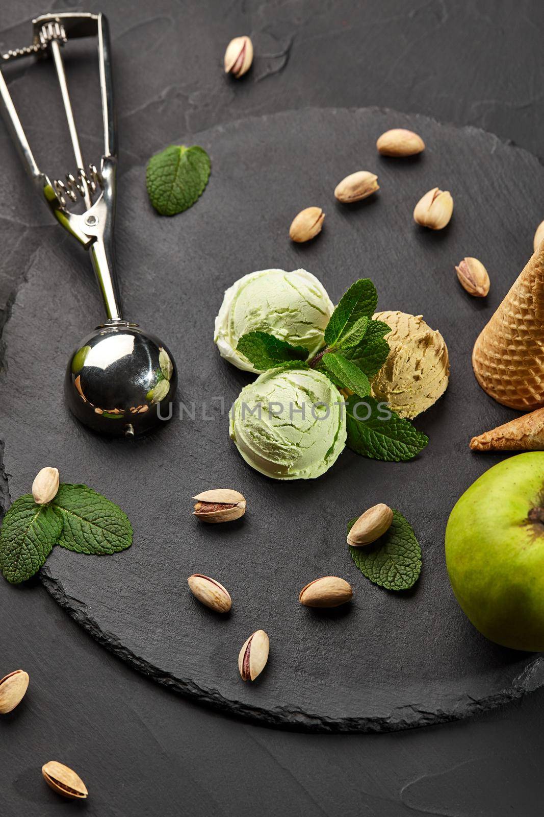 Close-up shot of a piquant pistachio and chocolate ice cream decorated with fresh mint, apple and scattered nuts, served on a stone slate over a black background. A metal scoop is laying nearby. Top view.