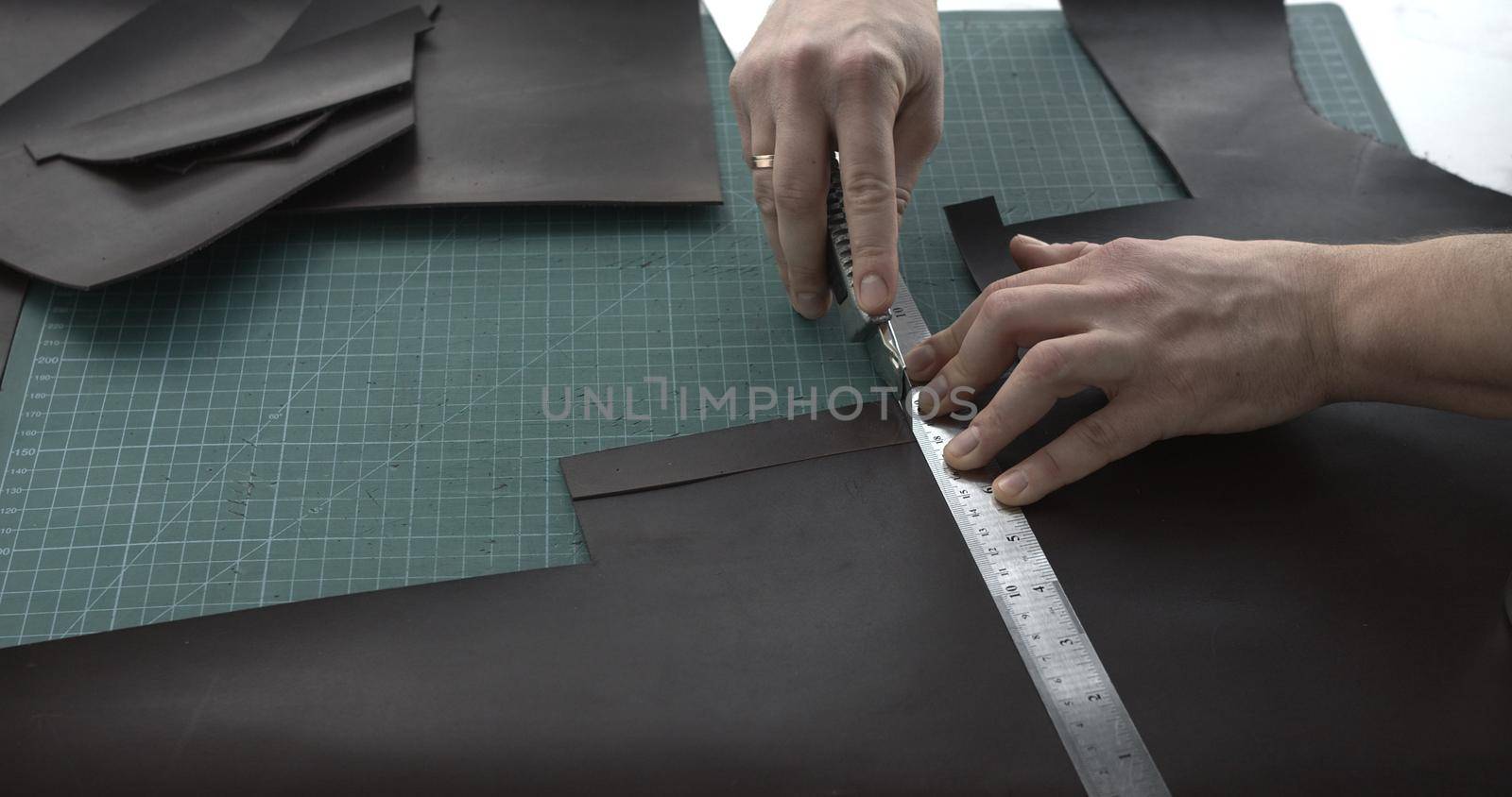 Close up of leather craftsman working with natural leather by knife and ruler by cutting a leather to a peaces. Handmade master at work in local workshop. Leather wallet stitch