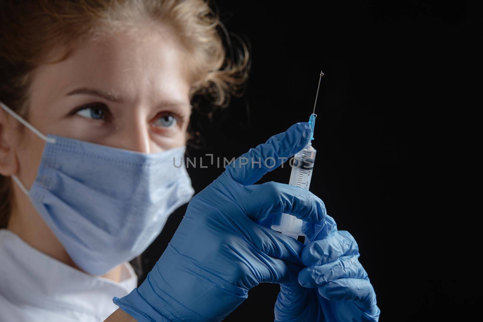 A female doctor wearing a protective mask and gloves holds a syringe with a vaccine or injection against a black background. A female nurse prepares to give an injection against a dark background. Woman with syringe in hand alone on black background. A drop of liquid on the syringe.