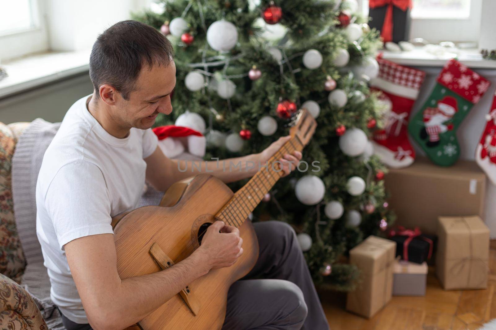 Happy young man is playing guitar. Guy is looking happily and carefree. Male in festive hat alone celebrating Christmas or new year. Christmas tree with garland in background.