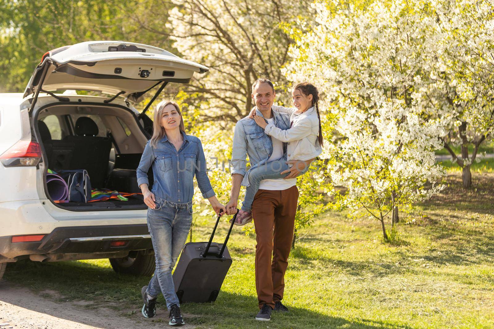 Happy family standing together near a car with open trunk enjoying view of rural landscape nature. Parents and their kid leaning on vehicle luggage compartment. Weekend travel and holidays concept. by Andelov13