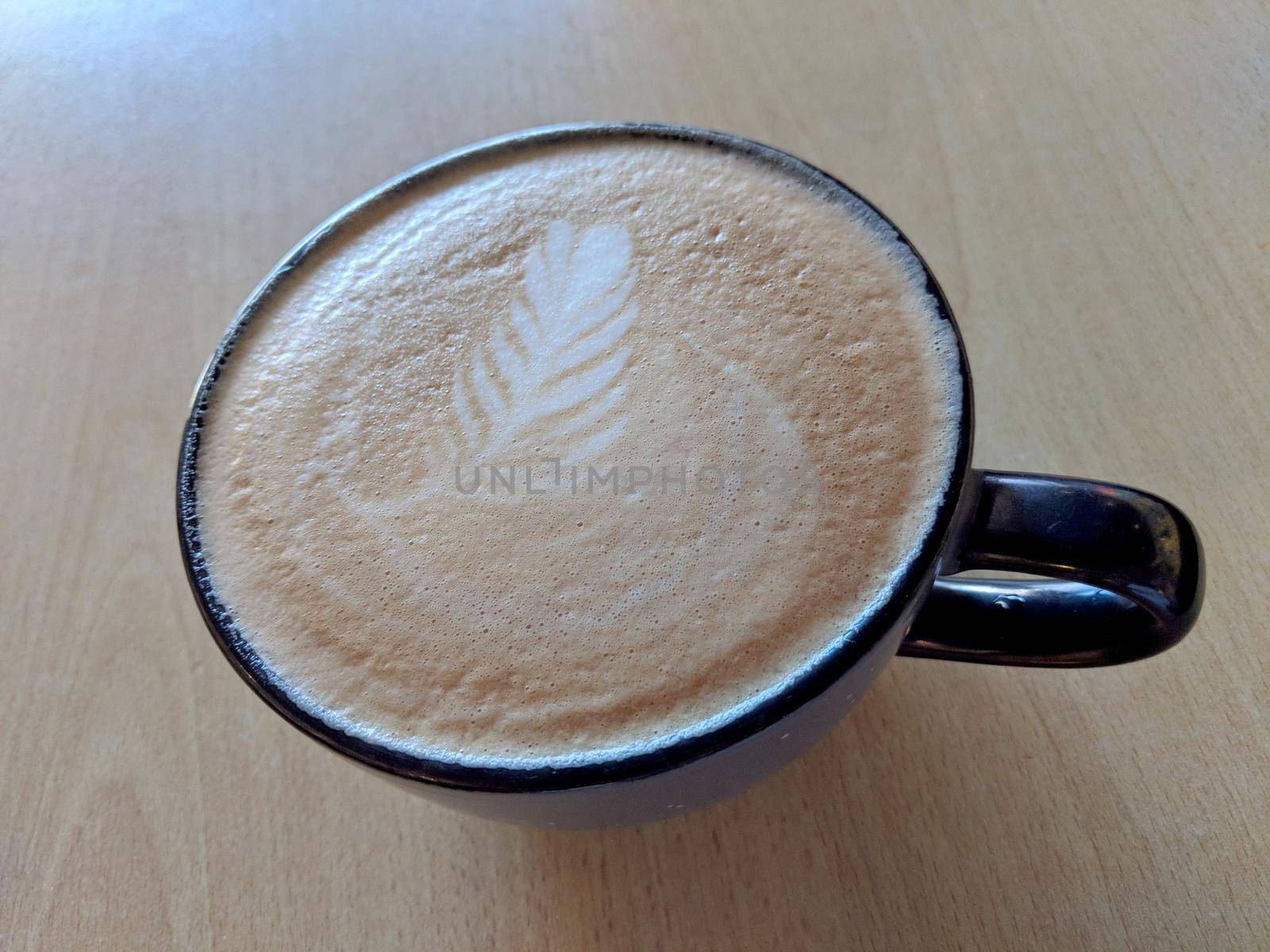 Balck cup of Cappuccino on saucer with a leaf pattern in foam on table.