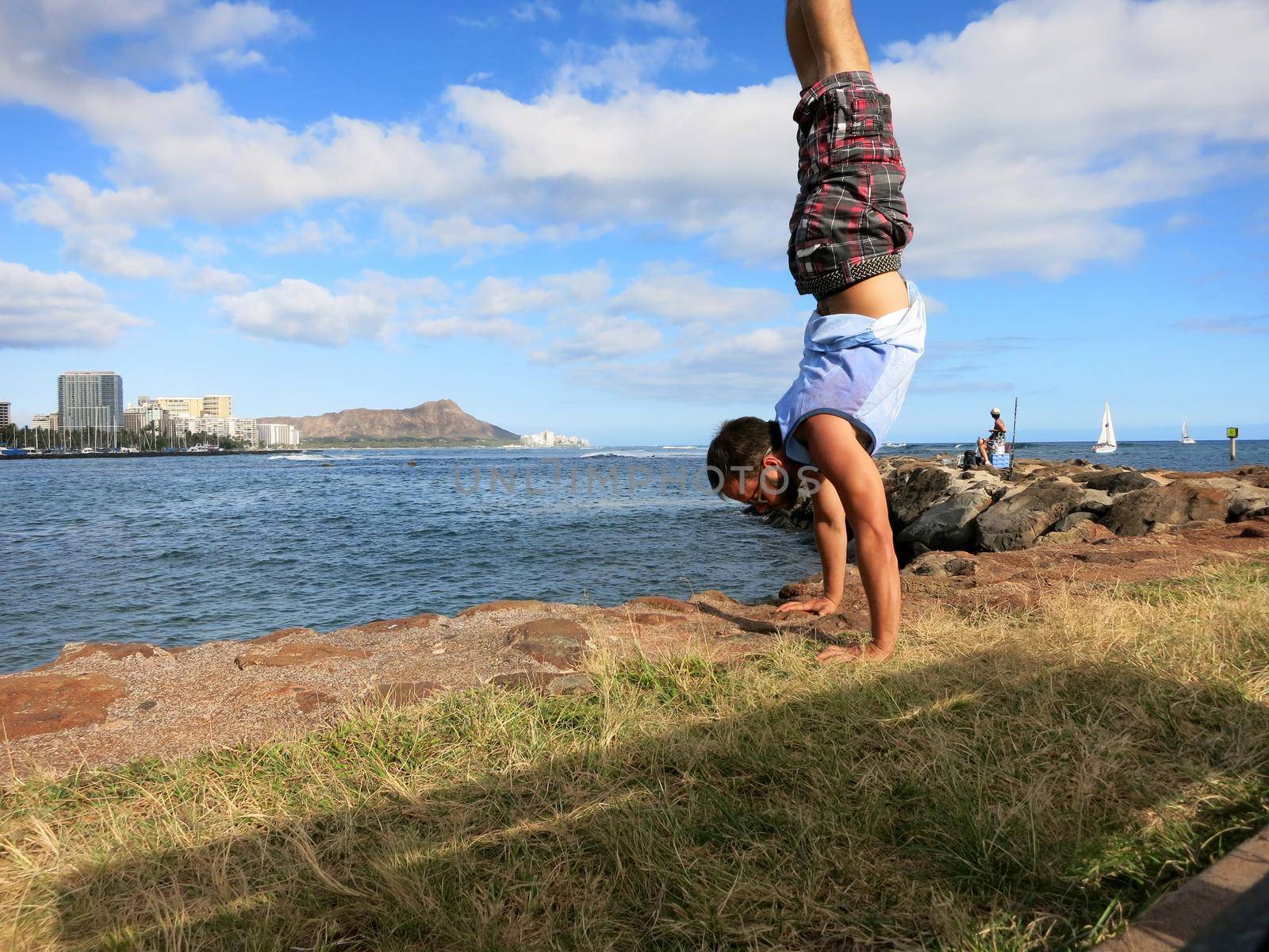 Man wearing shorts, shirt, and shoes Handstanding along shore of Magic Island with Ala Wai Boat Harbor, Waikiki, Diamond Head in the distance looking into the pacific ocean on Oahu, Hawaii on a beautiful day. 
