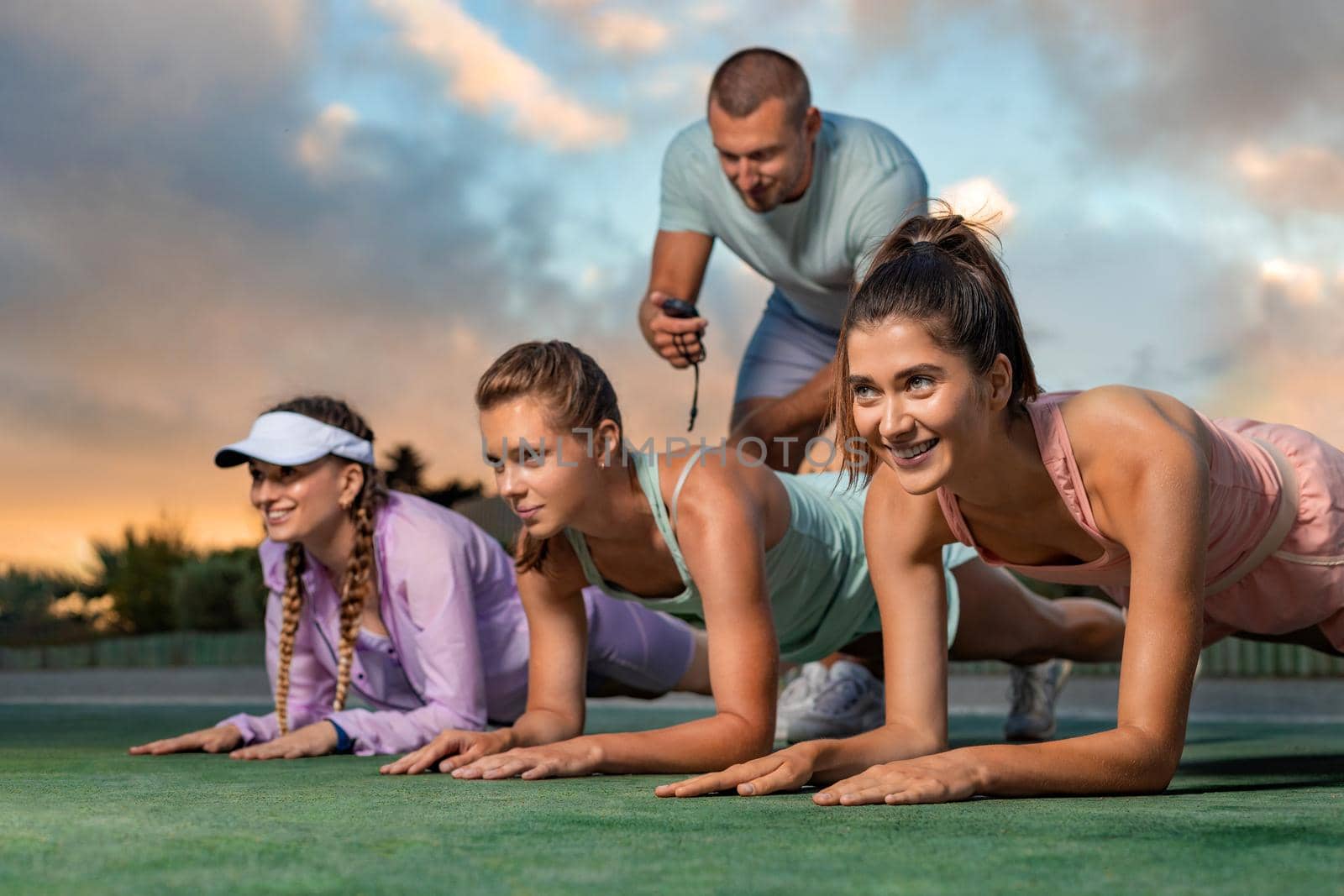 Team at plank excercises. Group of happy fitness women making body core planking exercise outdoors at gym. Male trainer with stopwatch. Healthy lifestyle motivation and sport concept. by MikeOrlov