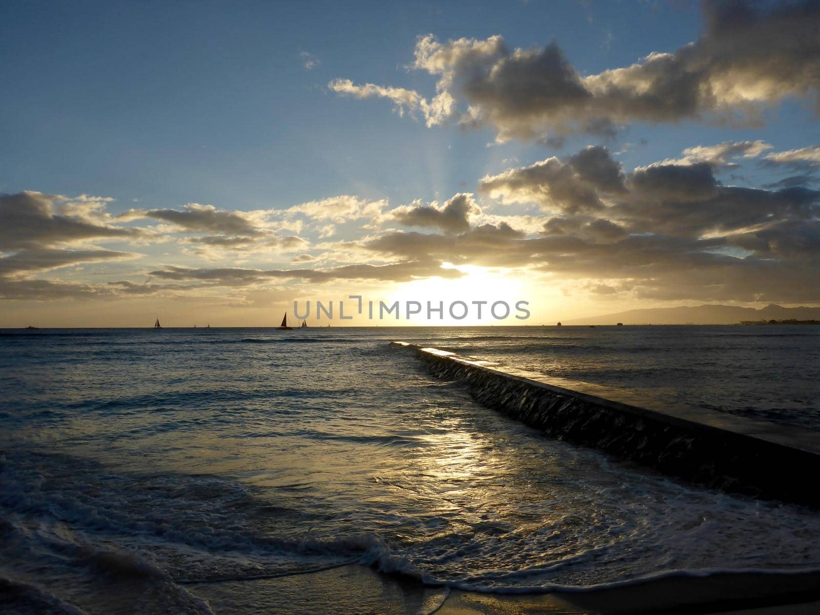 Rock Pier leads to Sunset over the Pacific ocean on the water with birds flying in the foreground and boats off the coast of Oahu, Hawaii with Waianae Mountain range visible. February 2016.